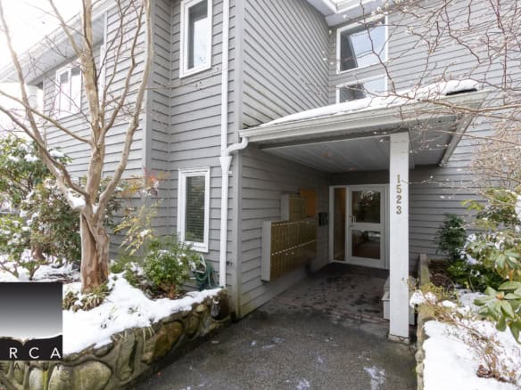 Cozy multi-family residential building entrance with snow-covered landscaping in Vancouver, BC. Modern grey siding, mailboxes, and inviting entryway showcase desirable Vancouver real estate.