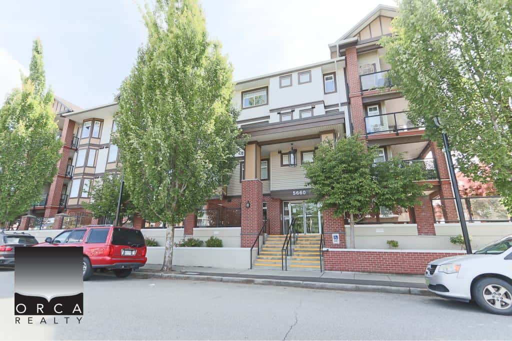 Bright multi-story residential apartment building with modern architecture, red brick accents, and large windows, surrounded by lush green trees and parked cars, located at 5660, in a vibrant neighbourhood.