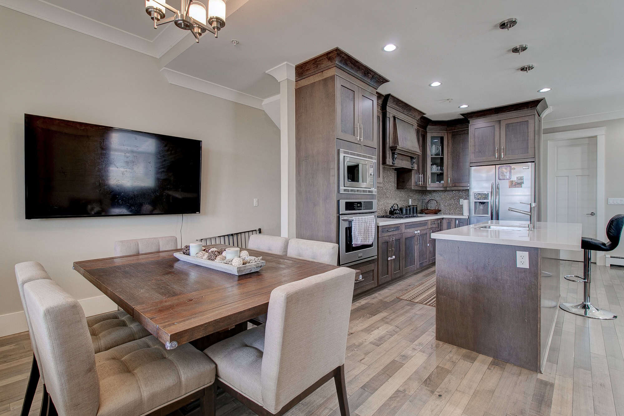 Modern kitchen and dining area with dark wood cabinetry, stainless steel appliances, island with breakfast bar, and a large wall-mounted flat-screen TV in a contemporary condo.