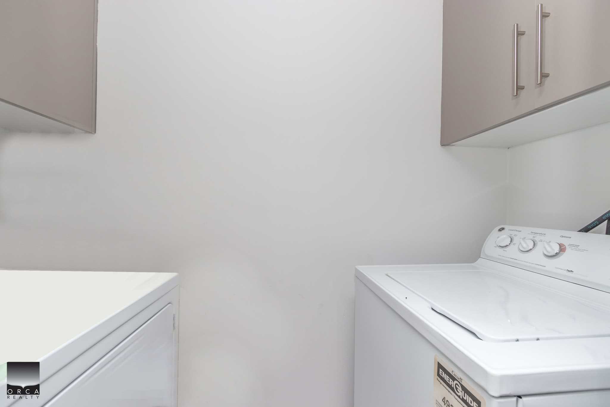 Bright laundry room with white washer and dryer, ample overhead storage cabinets, clean walls, perfect for a modern home in Victoria or Vancouver Island.