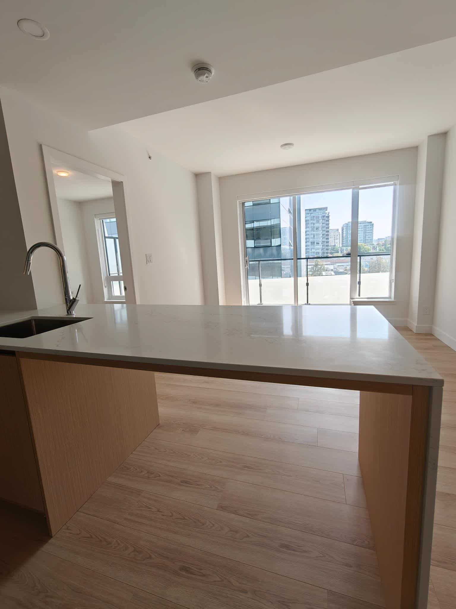 Bright modern apartment kitchen with view of city skyline, featuring white countertops, natural wood cabinetry, and large floor-to-ceiling windows, perfect for urban living in Vancouver.