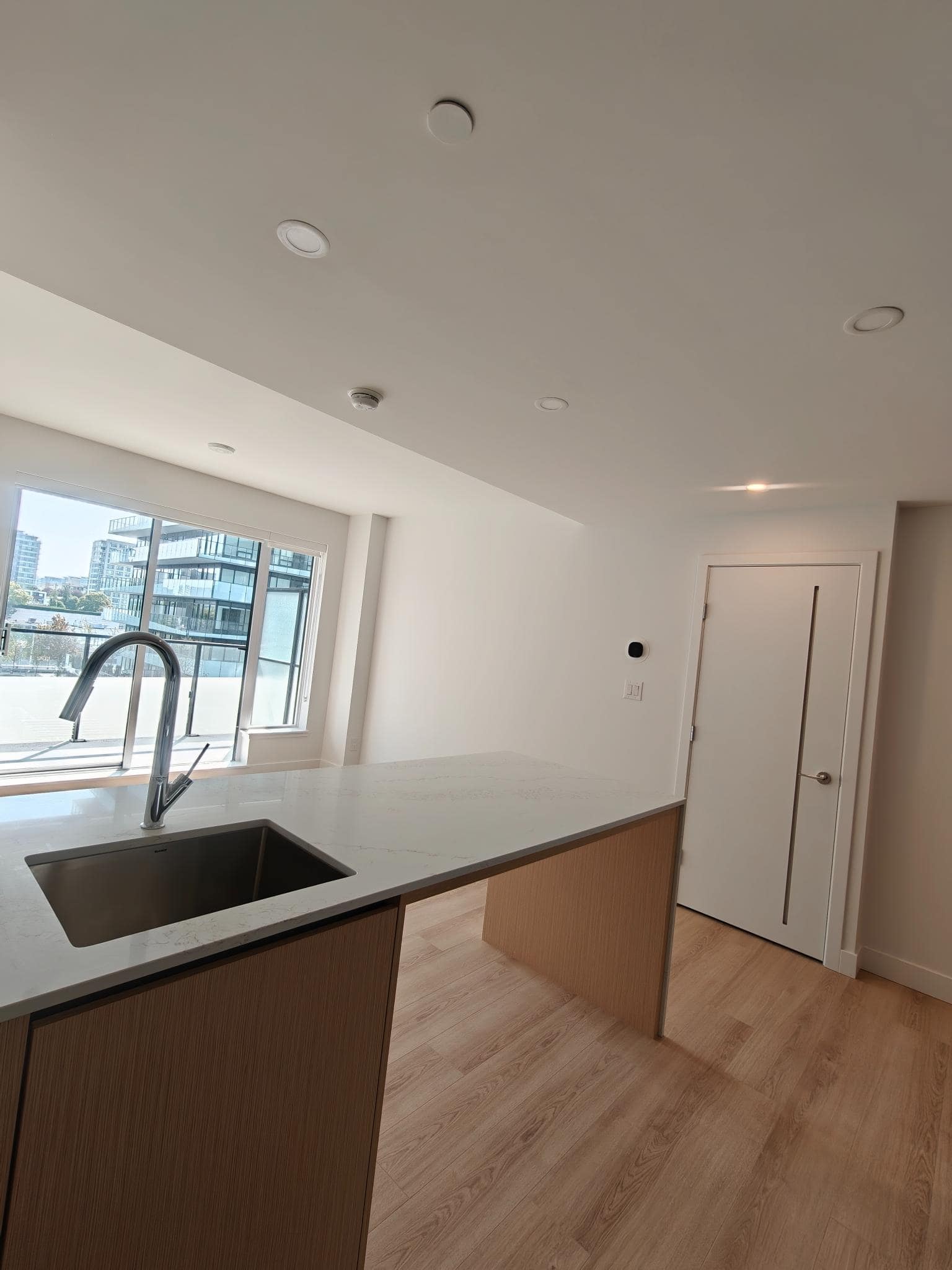 Bright modern kitchen in a new Vancouver condo, featuring a sleek white countertop, wood cabinetry, large window with city views, and contemporary lighting. Perfect space for stylish urban living.