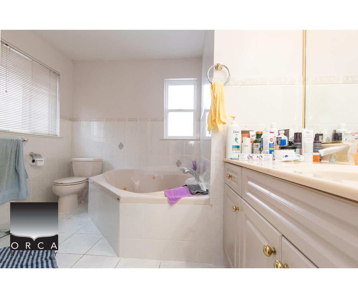 Spacious bathroom with a bathtub, toilet, and vanity, featuring ample natural light, white tile walls, and various personal care products, perfect for comfortable home living.