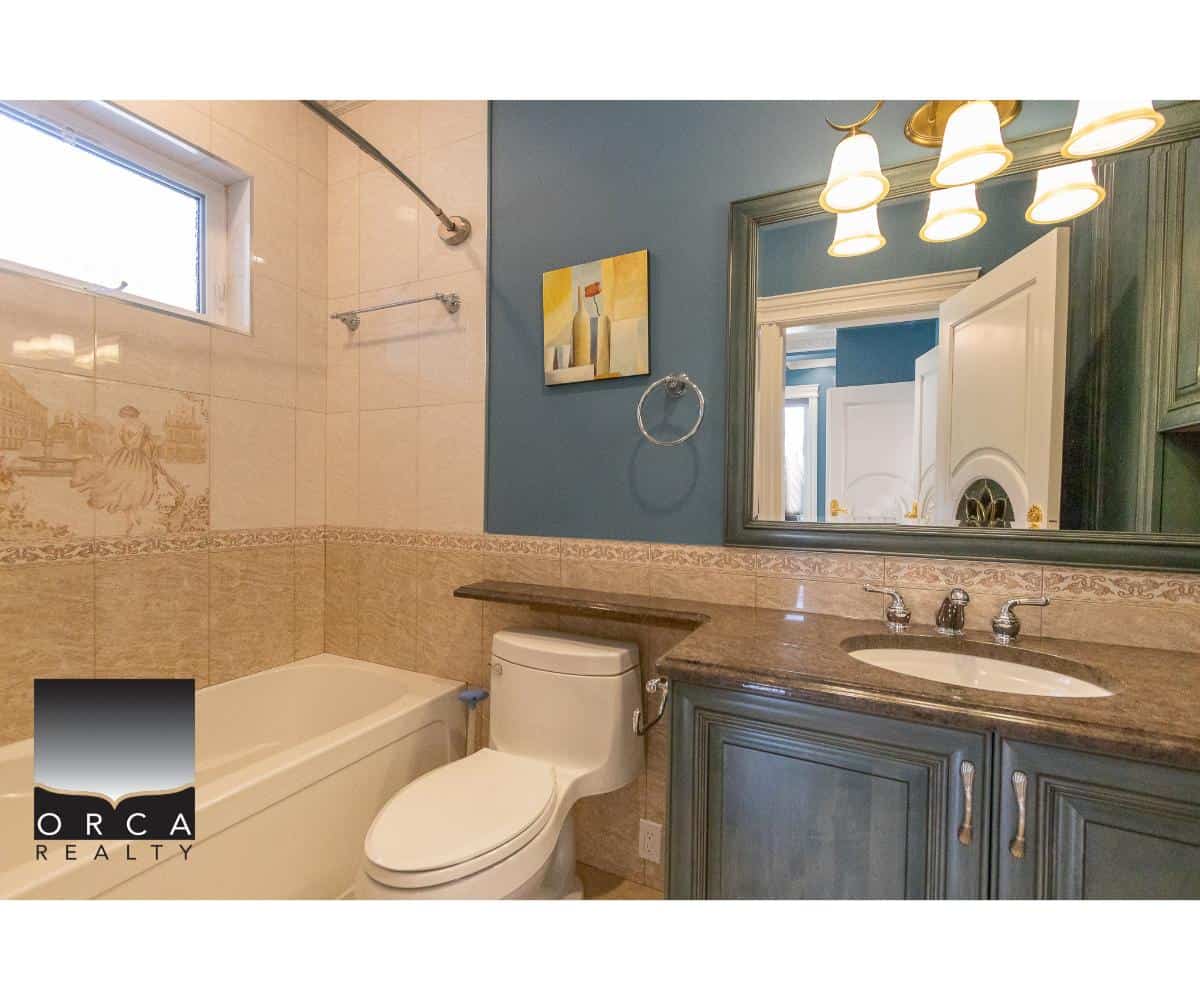Spacious bathroom featuring a bathtub, toilet, and a wooden vanity with a granite countertop, large mirror, and elegant lighting fixtures, showcasing interior design by Orca Realty Inc.