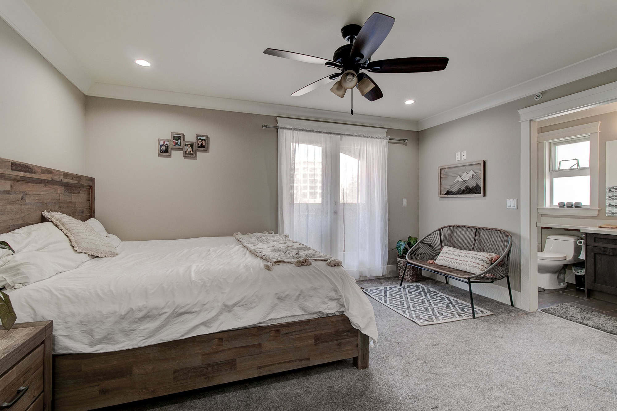 Spacious modern bedroom with a large wooden bed, ceiling fan, natural light from a sliding door with sheer curtains, cozy seating area, and an en-suite bathroom, perfect for Vancouver Island real estate.