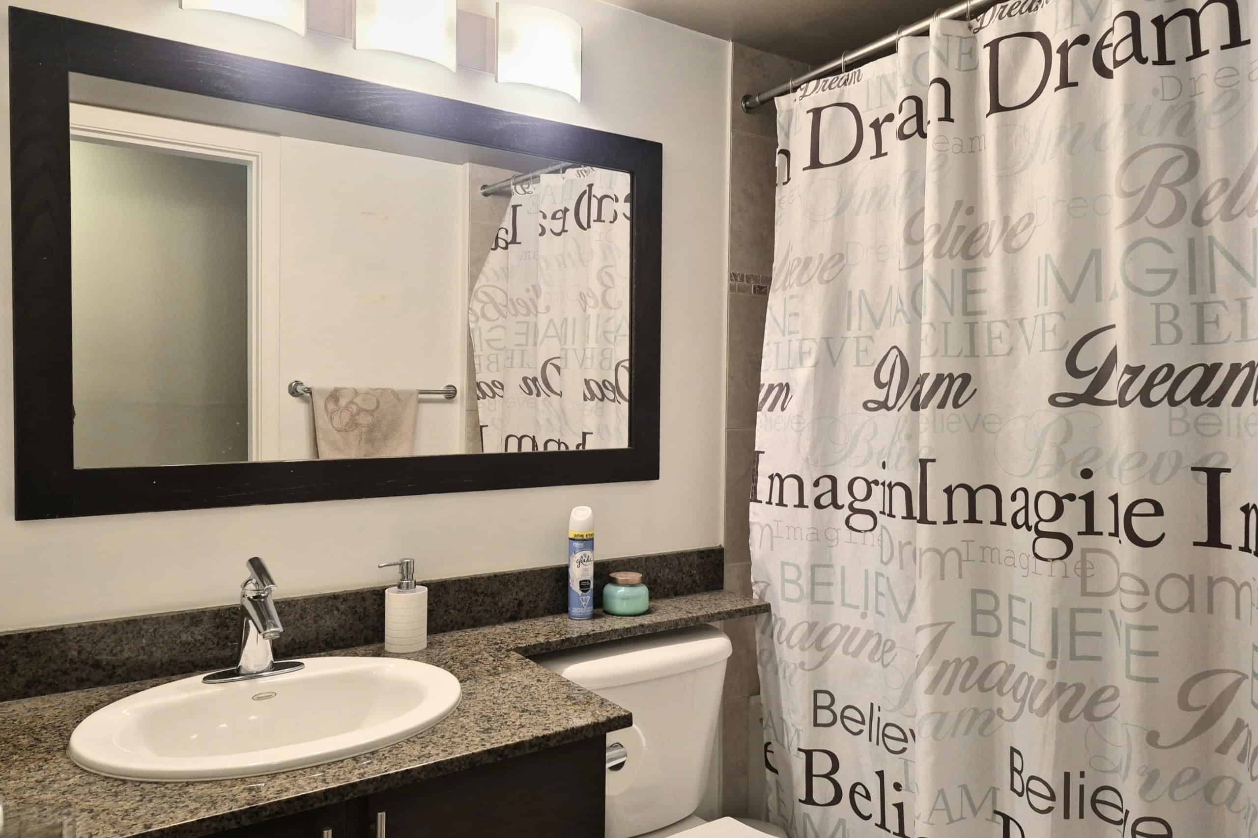 Modern bathroom interior featuring a granite countertop, oval sink, and large black-framed mirror, with a shower curtain decorated with inspirational words like "Dream," "Believe," and "Imagine," providing a stylish and motivational space.
