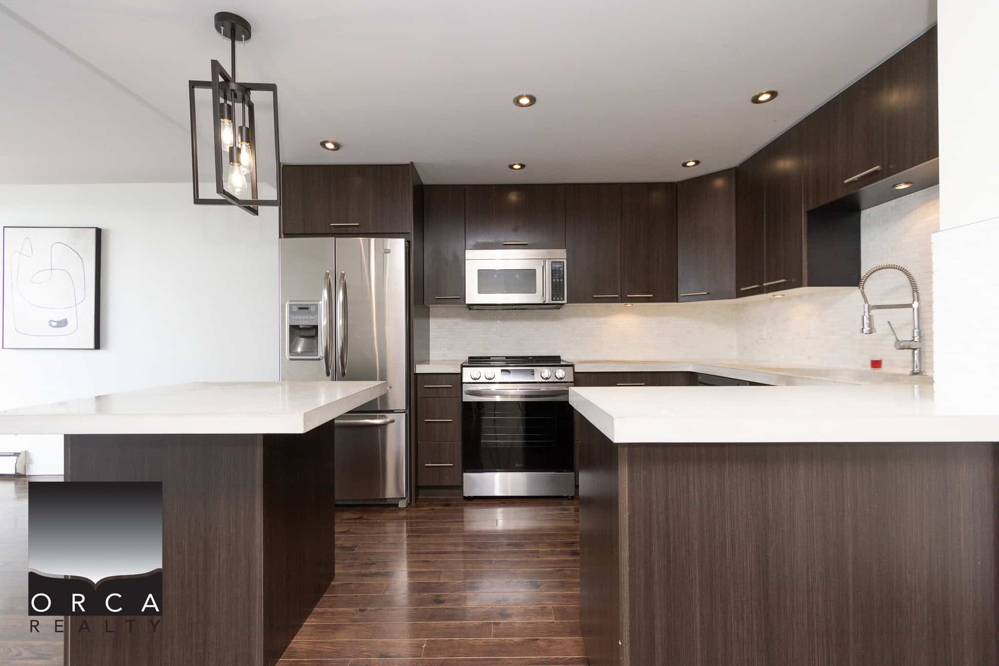 Modern kitchen with dark wood cabinets, stainless steel appliances, white countertops, and hardwood flooring, showcasing contemporary design and functionality.