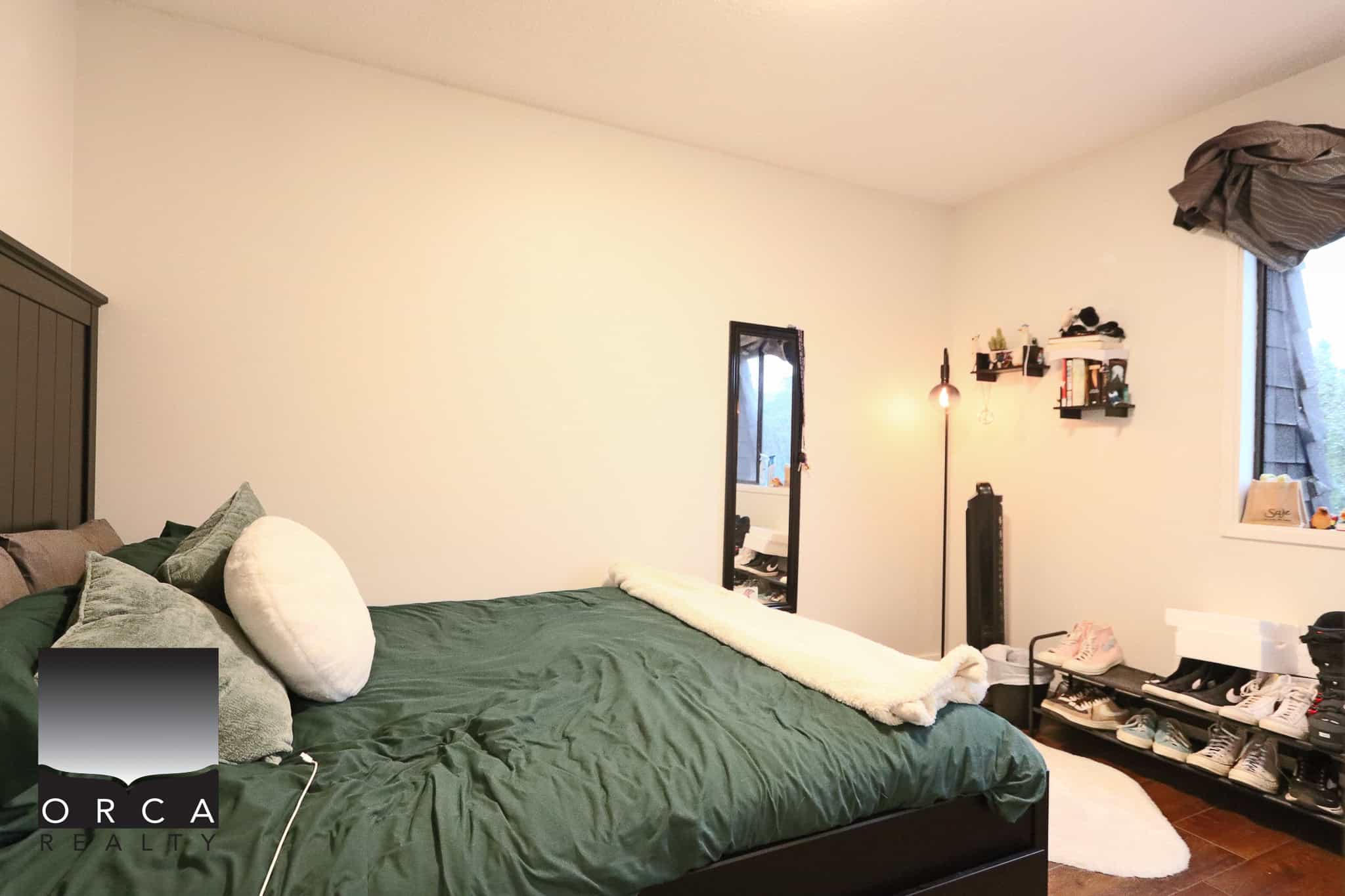 13 Cozy bedroom with a large bed, green bedding, and multiple plush pillows, featuring a full-length mirror, natural light from the window, and stylish shoe storage, ideal for Vancouver real estate listings.