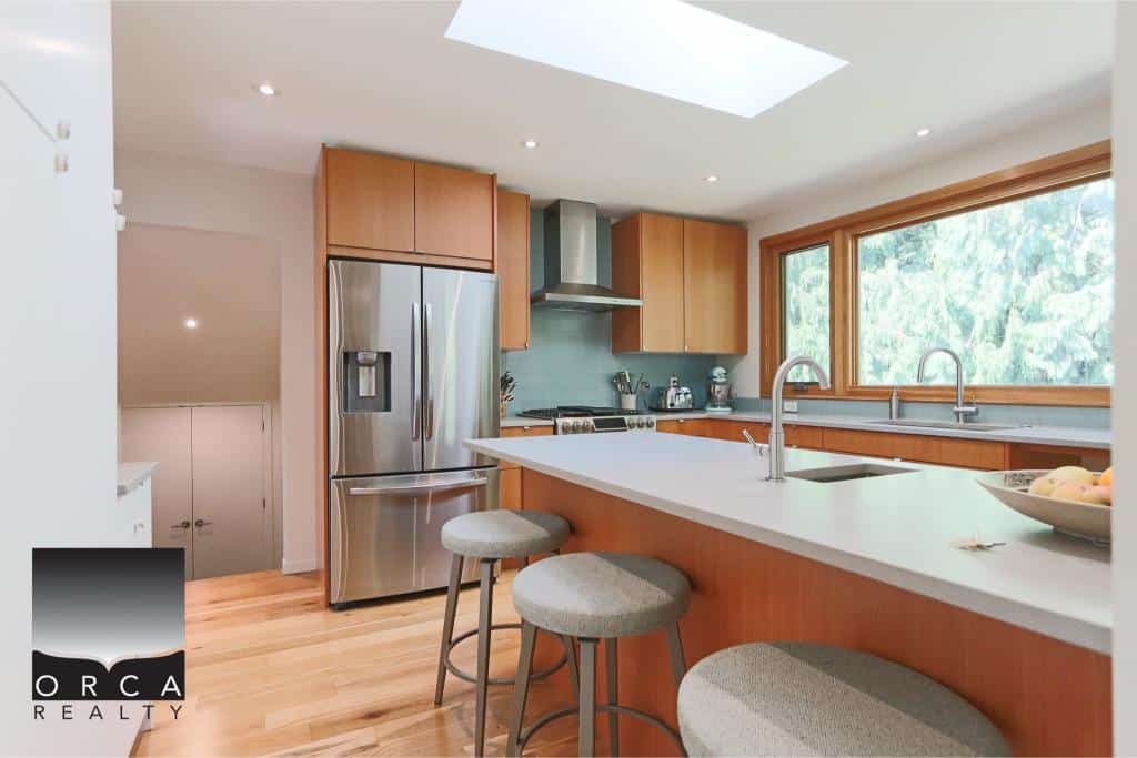 Stainless steel refrigerator and modern kitchen with ample cabinetry, natural light, and a large window showcasing greenery, perfect for Coastal BC living and real estate investments.
