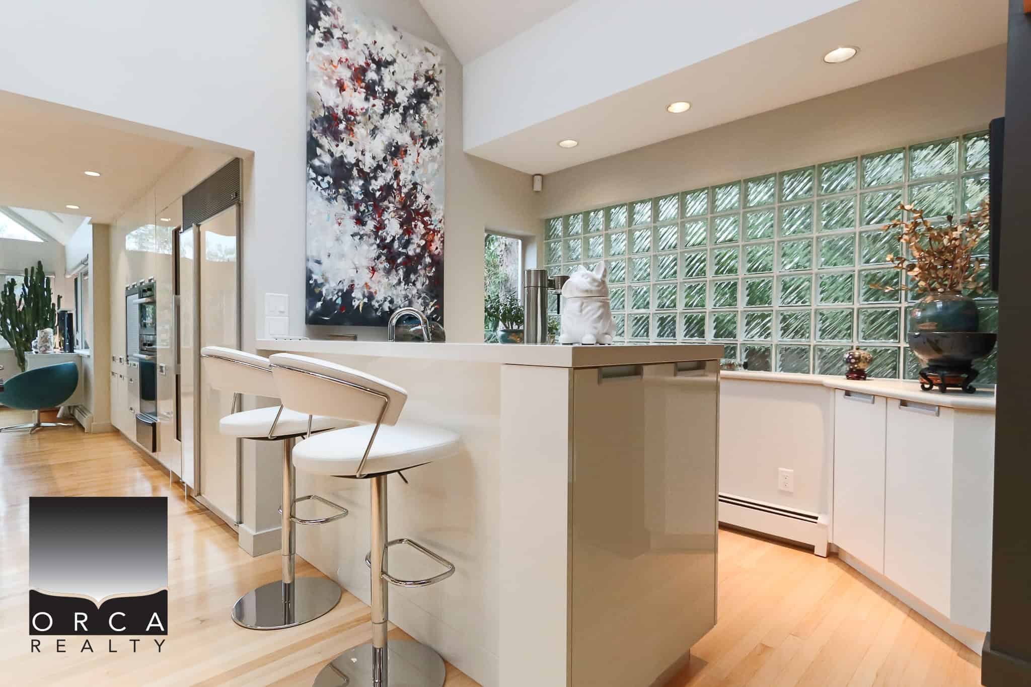 Bright modern kitchen with white cabinetry, a large island, artistic wall decor, glass block windows, and contemporary bar stools, showcasing elegant residential interior design in Victoria, BC.