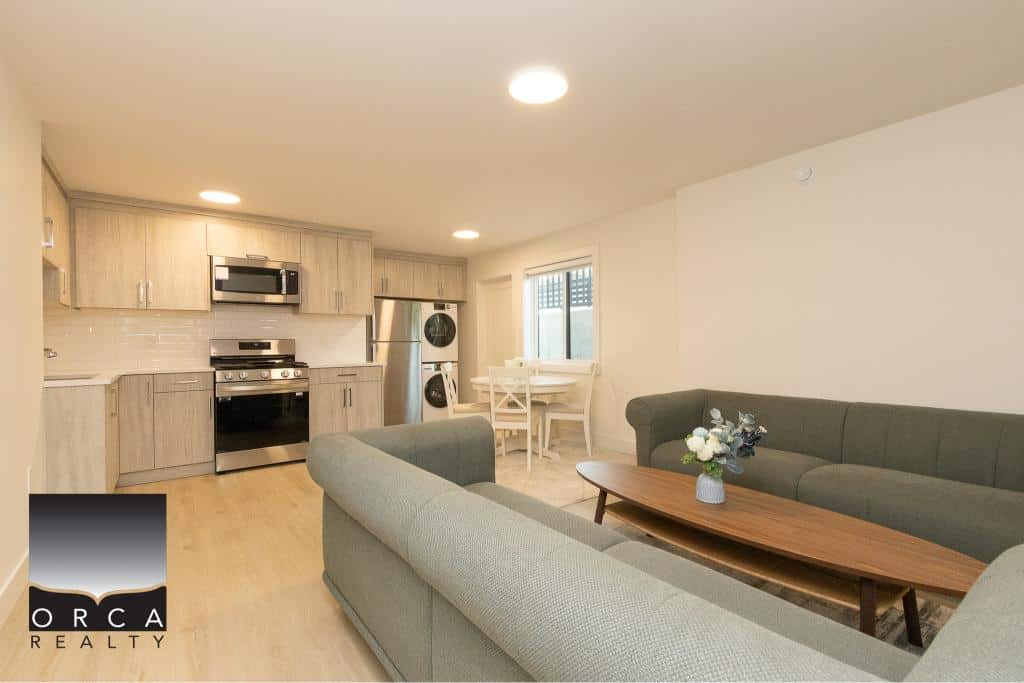Bright modern open-concept living room with kitchenette, featuring stylish neutral-toned sofas, contemporary kitchen appliances, and a cozy dining area in a Vancouver property listed by Orca Realty Inc.