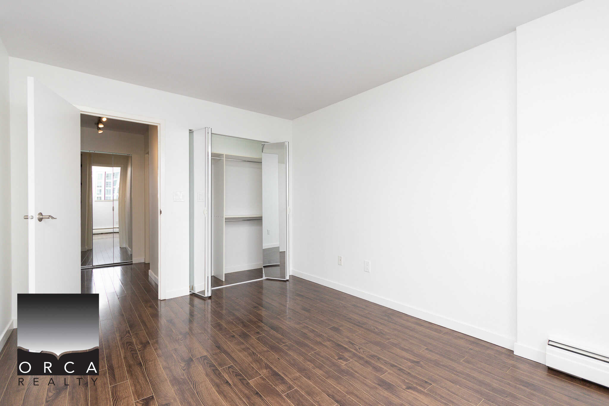 Bright, modern empty bedroom with dark hardwood floors, white walls, and a closet with mirrored bifold doors, ideal for Vancouver real estate buyers seeking contemporary condos or apartments.