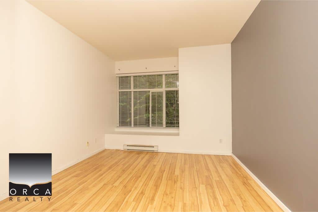 Bright empty living room with hardwood floors, large window with blinds, and neutral wall colors in a modern apartment.