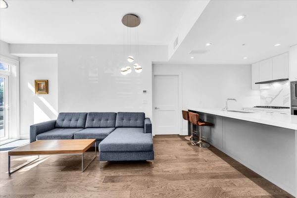 Spacious living room featuring a stylish blue sectional sofa, wooden coffee table, and natural light from large windows, located at 101-7878 Granville Street, Vancouver.