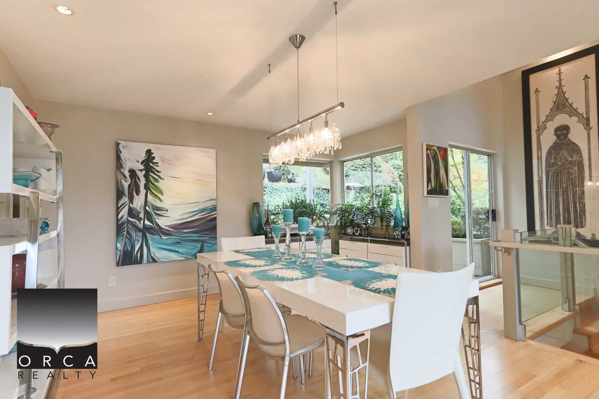 Modern dining room with contemporary decor, artwork, and large windows, featuring a sleek white dining table with turquoise accents, perfect for family meals and entertaining, by Orca Realty Inc.