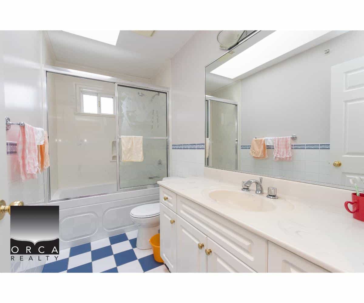 Bright bathroom with a white vanity, large mirror, shower/tub combo with sliding glass door, colourful towels, and a checkered blue and white floor, showcasing modern home features by Orca Realty Inc.