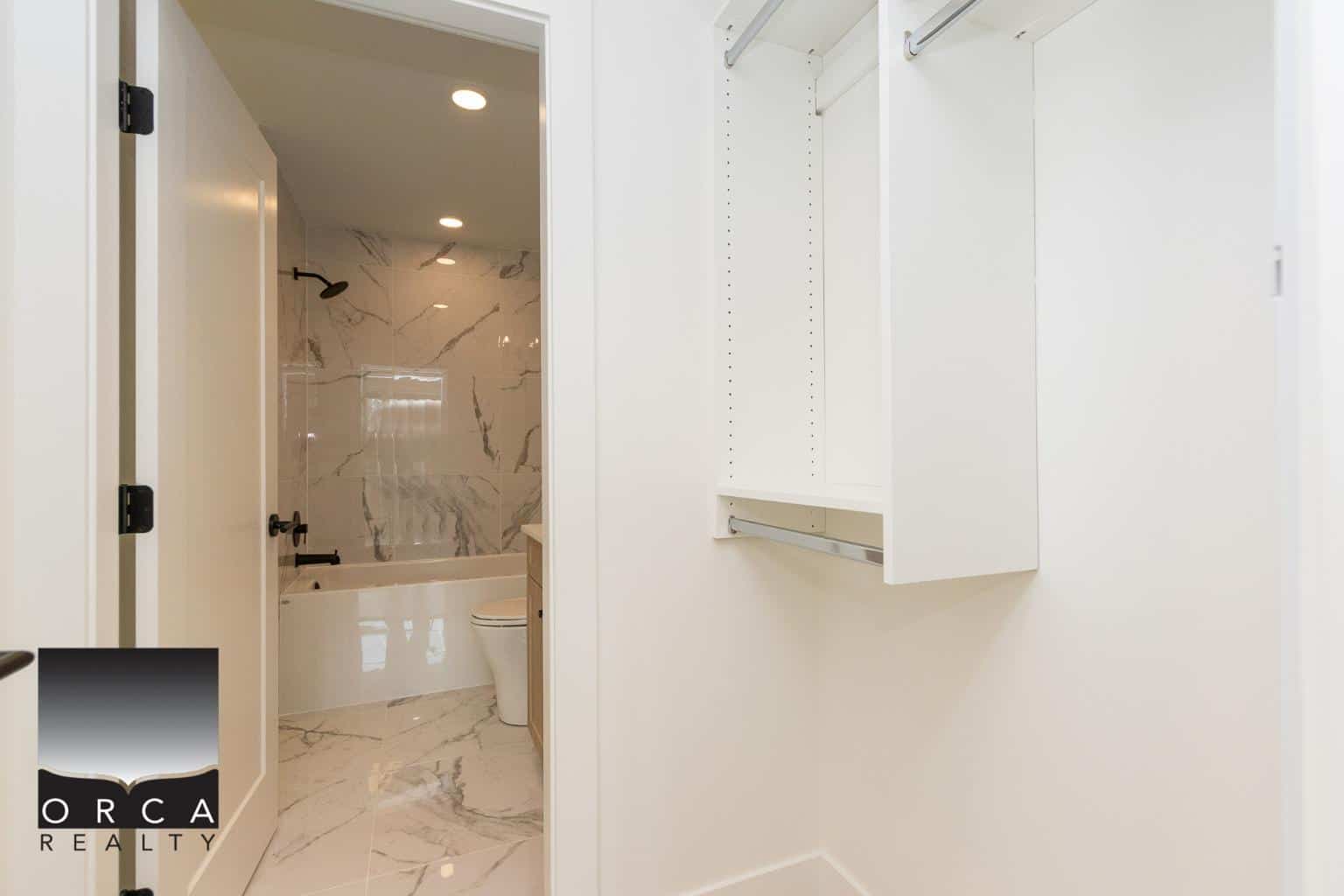 Modern walk-in closet with built-in shelving and hanging space in a new home listed by Orca Realty Inc., with a stylish bathroom featuring marble tiles in background.
