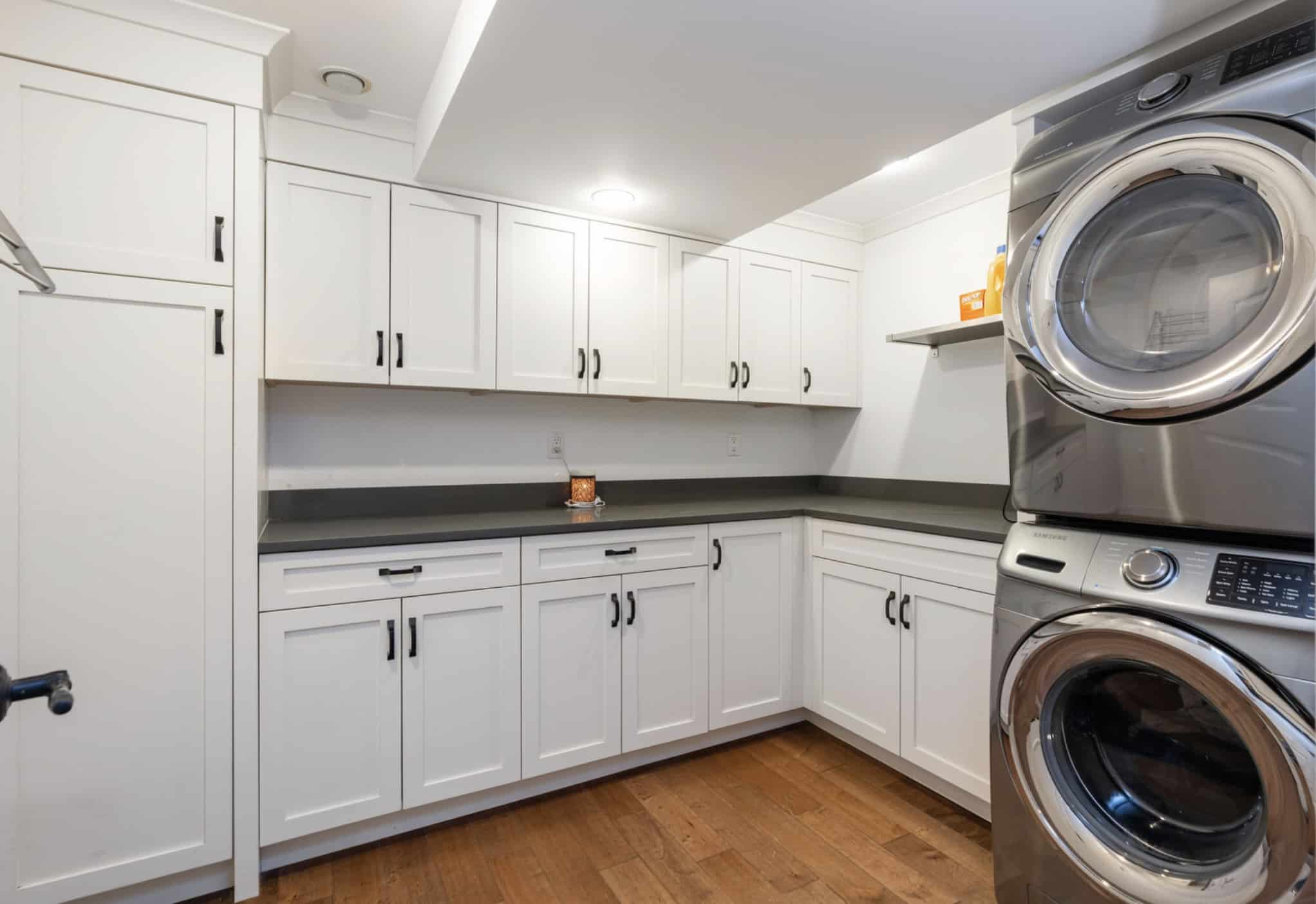 Screenshot Modern laundry room with stackable washer and dryer, white cabinetry, grey countertops, and hardwood flooring, showcasing efficient home organization and real estate features by Orca Realty Inc.