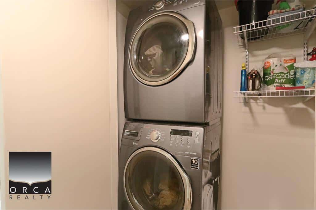 Stacked front-loading washer and dryer in a laundry room with shelving for supplies, showcasing home appliances from Orca Realty Inc.