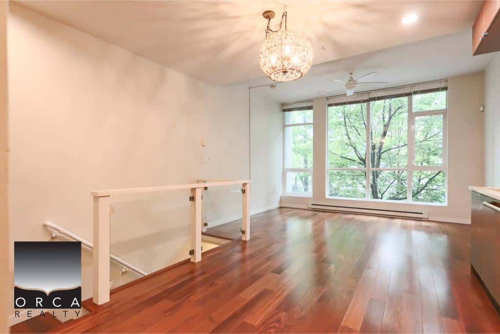 Bright spacious living area with large windows overlooking trees, hardwood floors, modern kitchen, and elegant chandelier, ideal for comfortable BC home living.