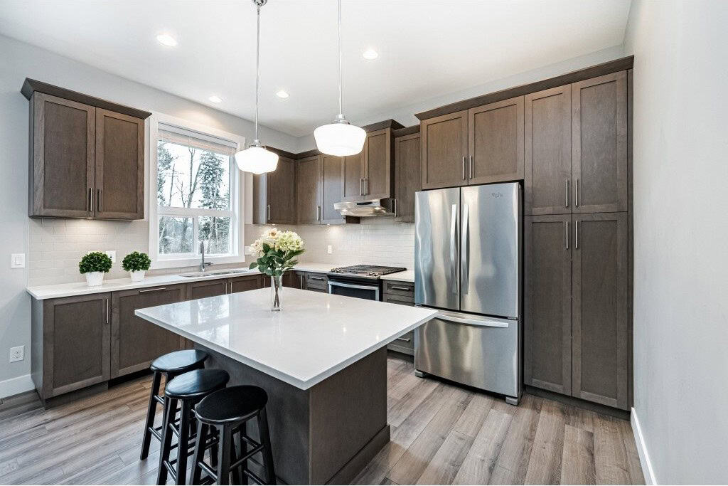 Modern kitchen with grey cabinets and stainless steel appliances, including a refrigerator and stove, featuring a central island with a white countertop, pendant lighting, and natural light from a window.