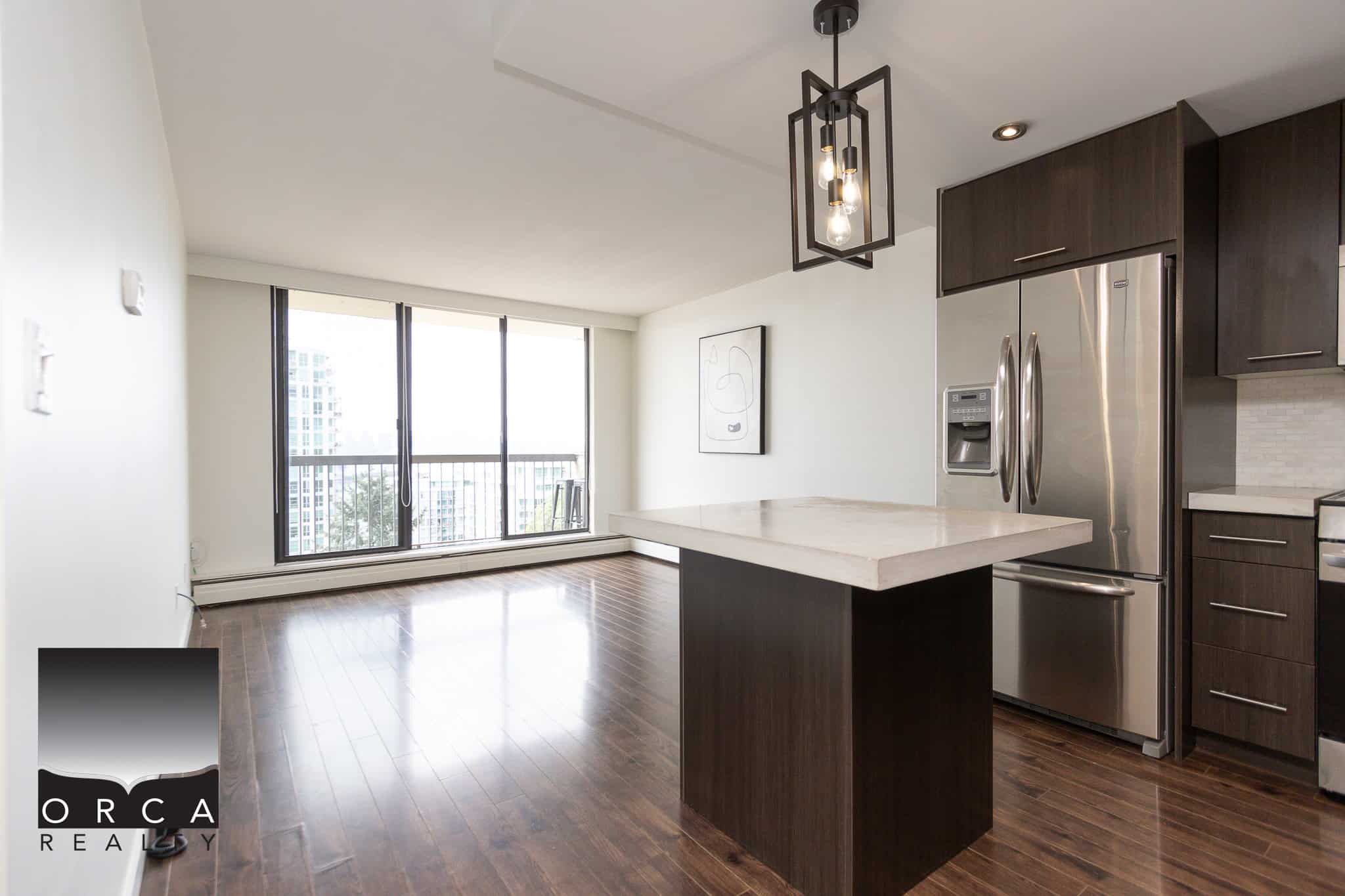 Modern open-concept kitchen and living space in downtown Vancouver, featuring stainless steel appliances, sleek dark wood cabinets, hardwood floors, and floor-to-ceiling windows with city views.