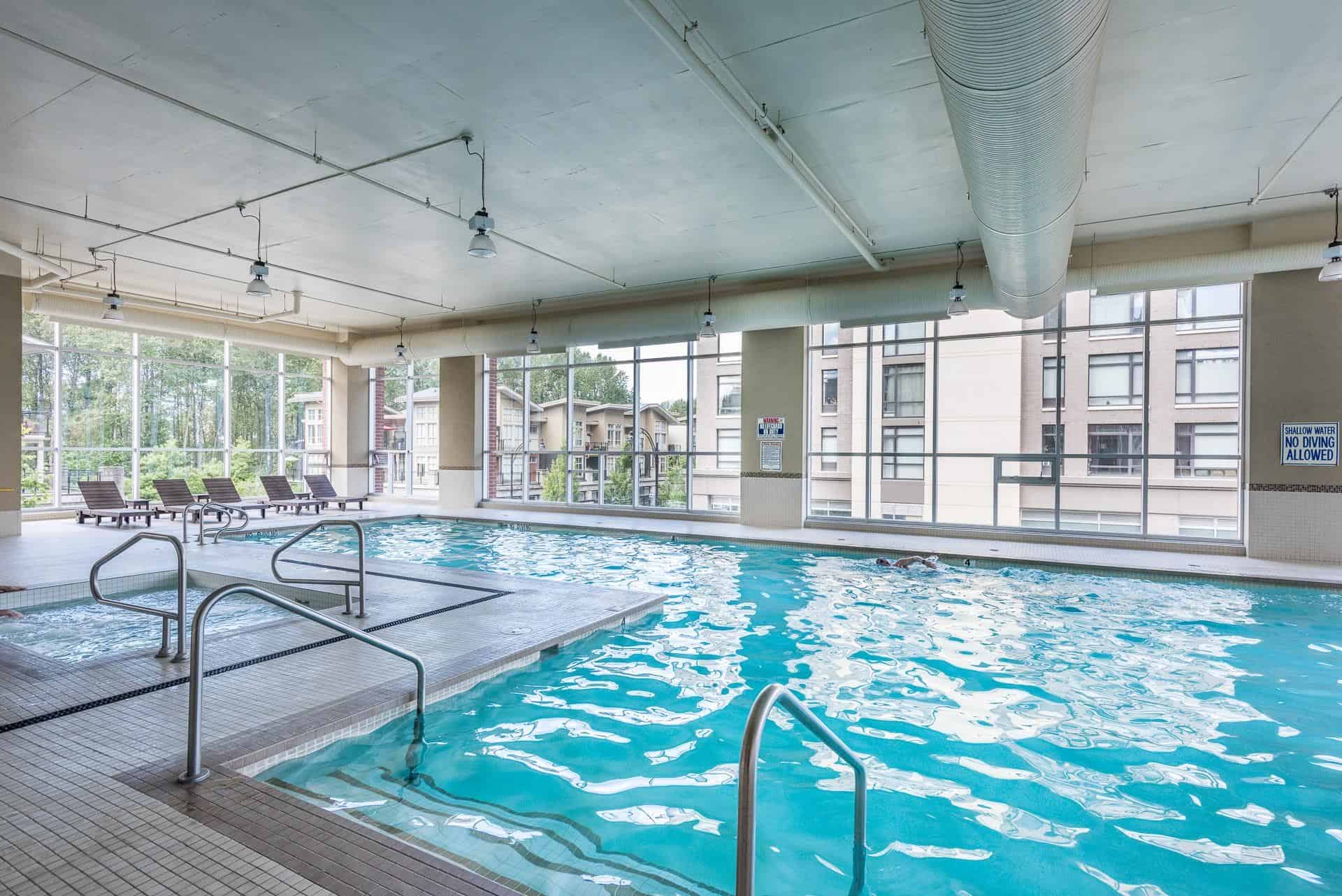 Modern indoor swimming pool at Orca Realty Inc. with large windows, lounge chairs, and clear water, ideal for relaxation and fitness in Vancouver's vibrant real estate community.