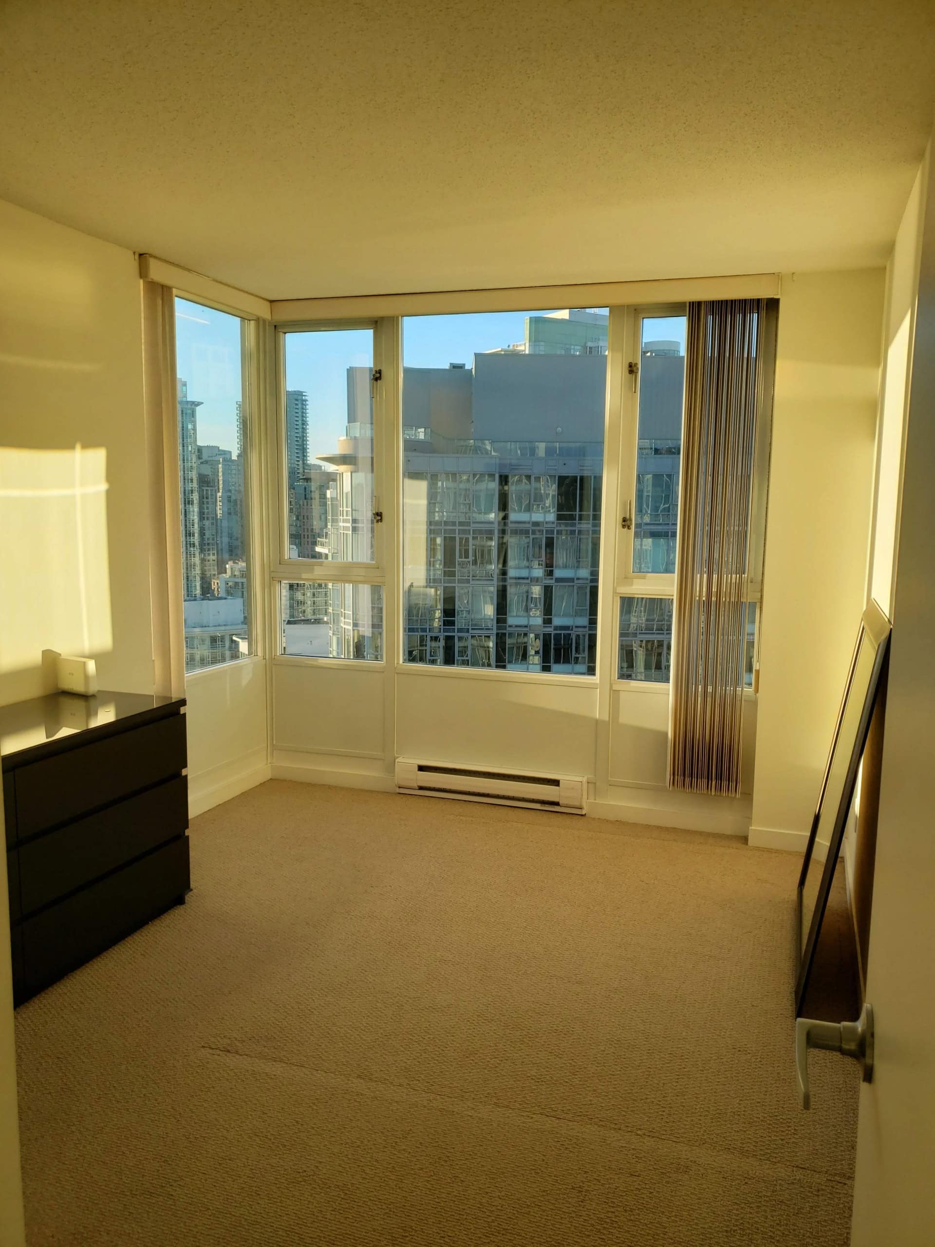 Modern high-rise condo bedroom with large windows offering city skyline views, beige carpet, and minimal furniture. Ideal for urban living in downtown Vancouver near amenities.