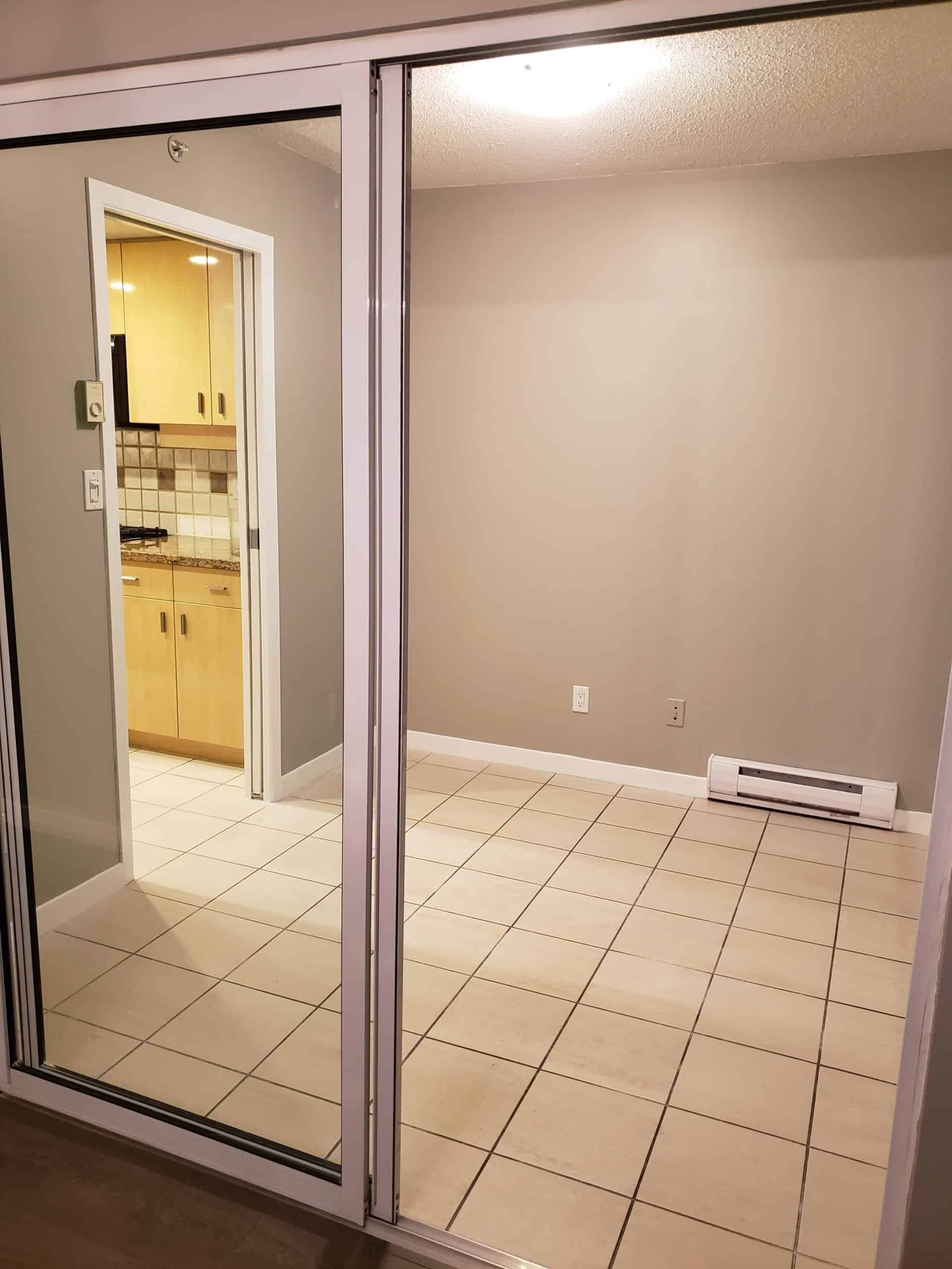 Modern apartment interior with a sliding glass door, beige tiled flooring, neutral wall paint, and a view into a compact kitchen, ideal for Vancouver condo living and real estate investment.