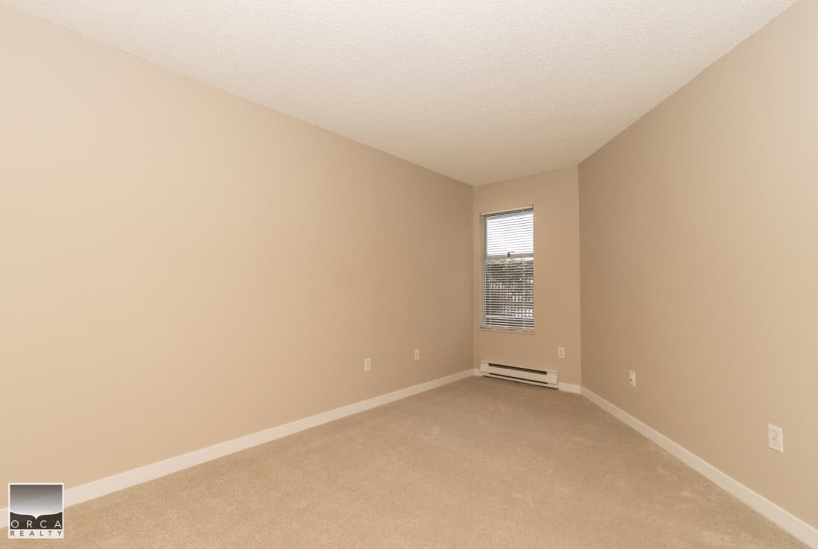 Bright empty bedroom with beige walls, carpet flooring, a window with blinds, and baseboard heating, ideal for Vancouver property listings by Orca Realty Inc.