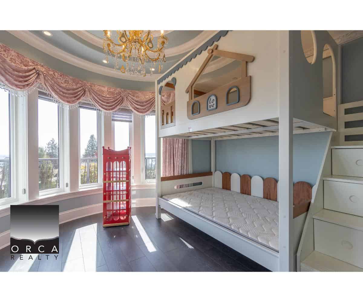 Bright children's bedroom with a custom bunk bed, large bay windows, elegant pink drapes, and a chandelier, showcasing beautiful interior design by Orca Realty Inc. in BC.