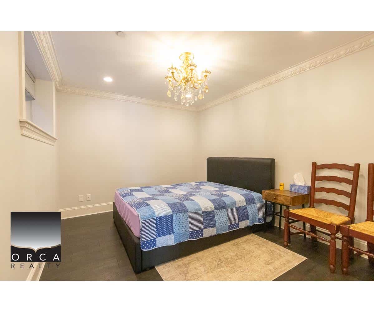 Bedroom with a black upholstered bed frame, blue and white checkered bedding, wooden chairs, small side table, chandelier, and neutral wall paint in a modern home interior.