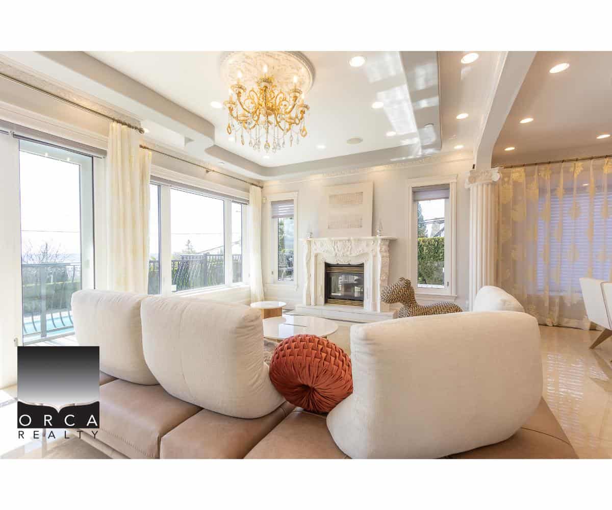Luxurious living room with large windows, elegant chandelier, white marble fireplace, and stylish furniture, showcasing upscale real estate in Victoria BC by Orca Realty Inc.