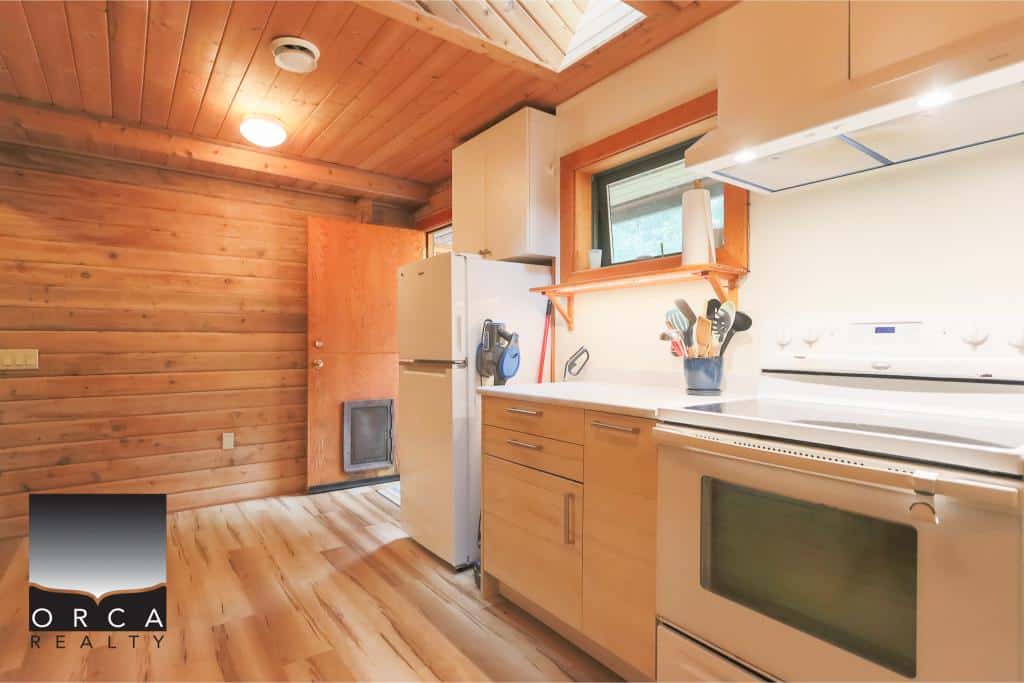 Cozy wooden kitchen in a cabin-style home with white appliances, natural wood cabinets, and a small window, showcasing warm interior design and functional kitchen space in Orca Realty listing.