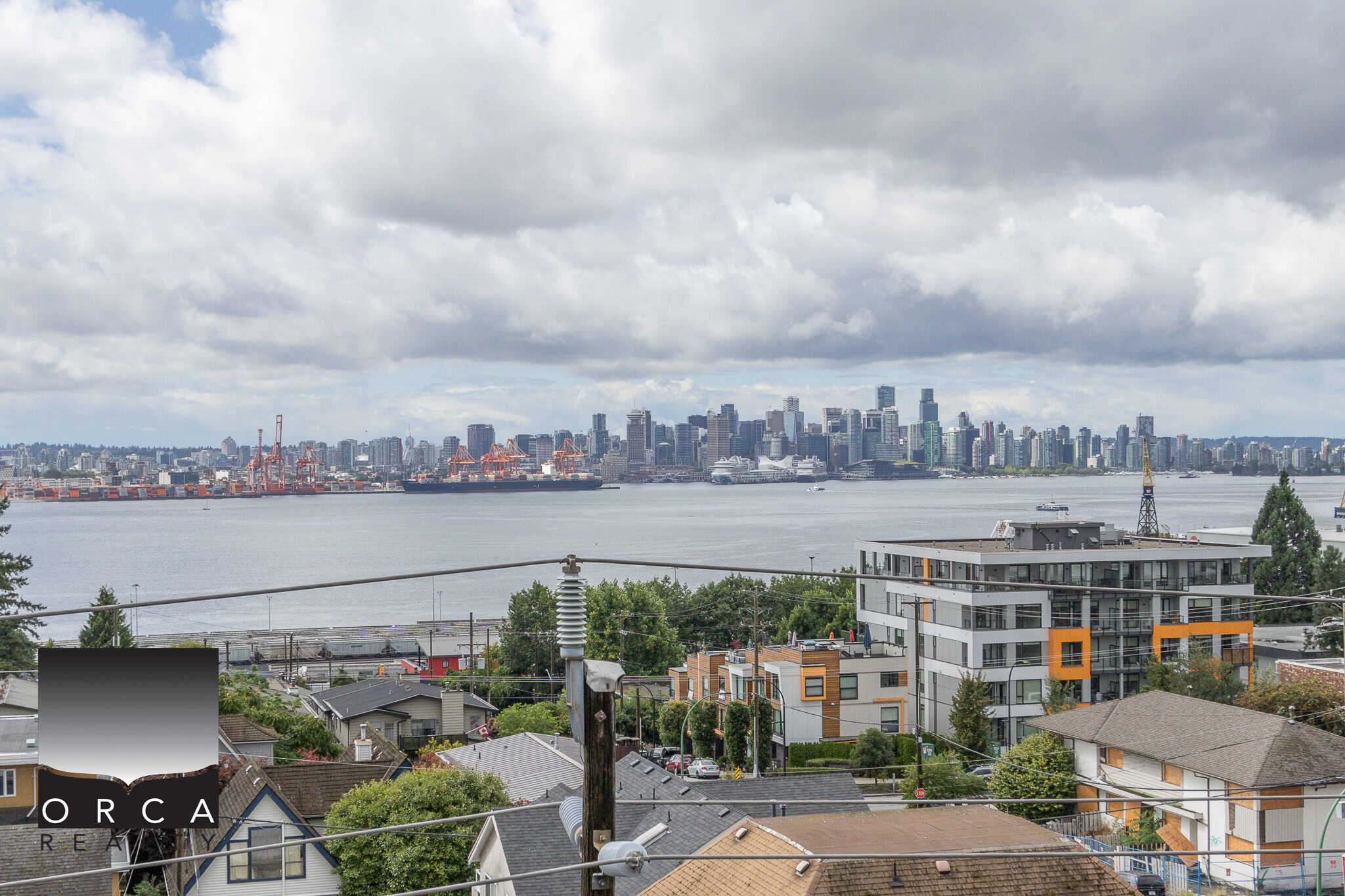 Panoramic view of Vancouver skyline and waterfront from a residential neighbourhood in North Vancouver, showcasing modern condos, rooftops, and lush greenery.