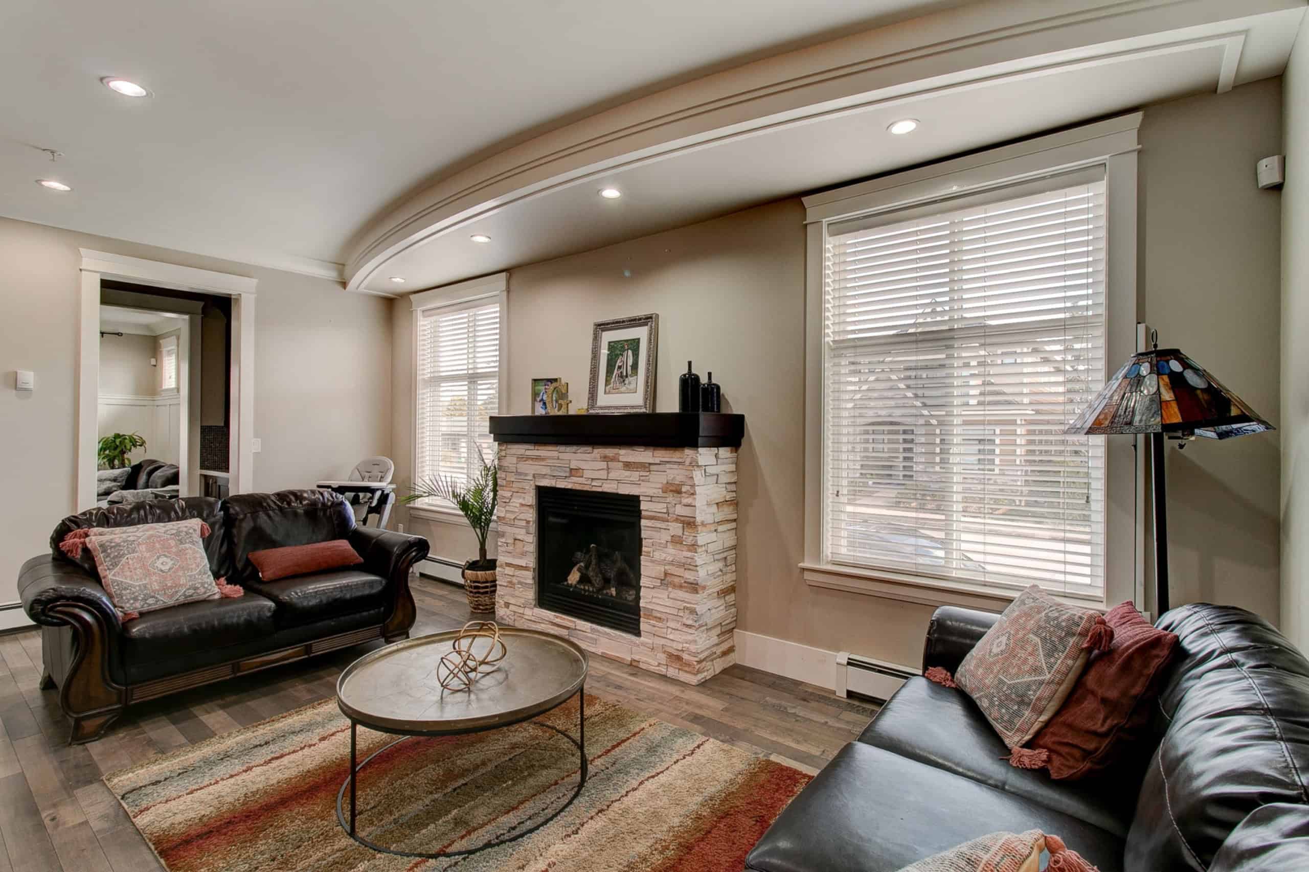 Spacious living room with black leather sofas, a stone fireplace, and large windows with white blinds, showcasing a cozy, modern interior design perfect for family gatherings and entertaining.