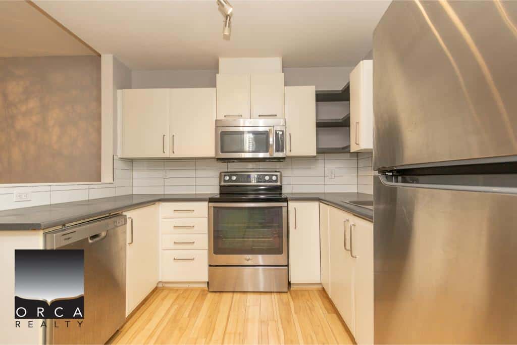 Modern kitchen with stainless steel appliances including a refrigerator, microwave, and oven, featuring white cabinetry and wooden flooring. Perfect for Vancouver Island homebuyers seeking a stylish, functional kitchen.