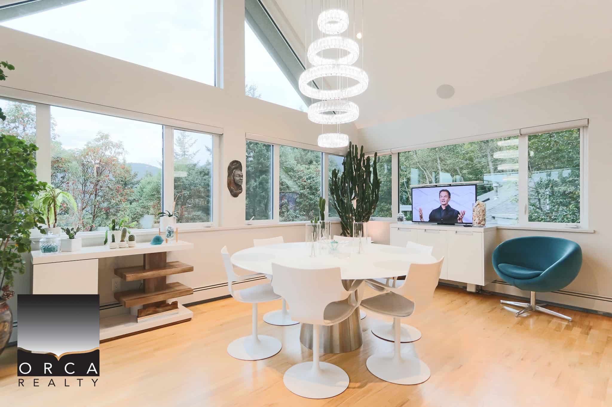Modern sunlit dining room with large windows, green outdoor views, stylish interior decor, and contemporary furniture, ideal for luxury Vancouver Island homes.