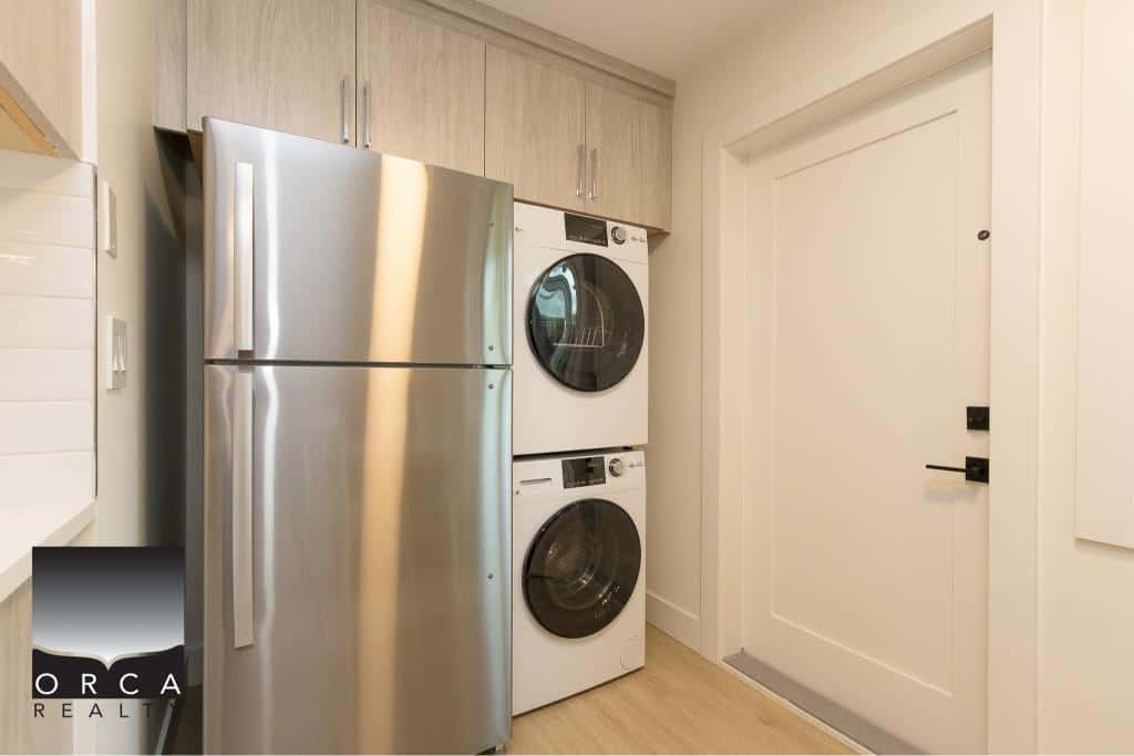 Stainless steel refrigerator with washer and dryer stacked in a modern laundry area at Orca Realty Inc., highlighting contemporary appliances and efficient space utilization for residential listings.
