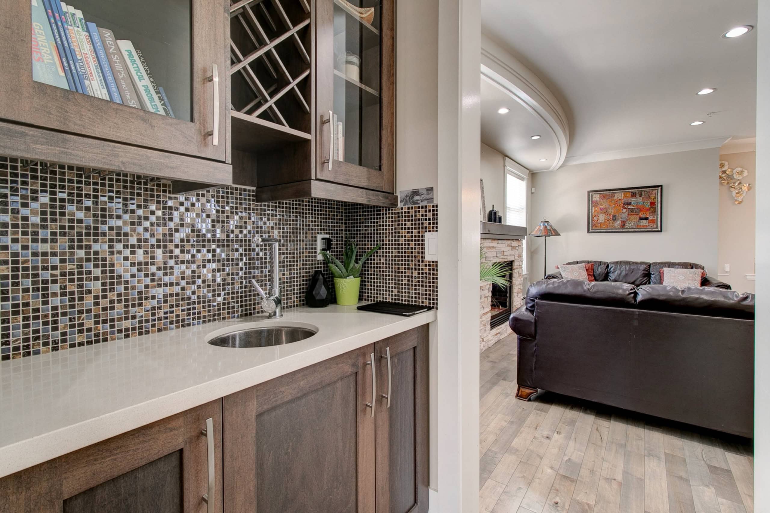 Modern home wet bar with wood cabinets, mosaic tile backsplash, and integrated wine storage, seamlessly connected to a cozy living room with fireplace, offering stylish and functional entertainment space.