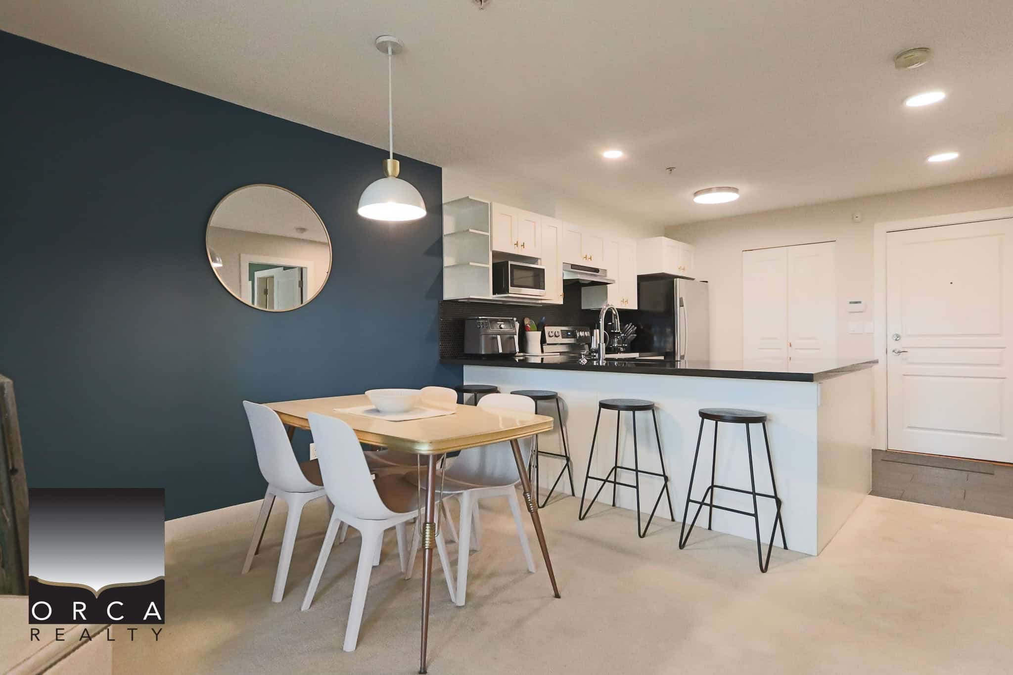 Modern kitchen and dining area inside a contemporary condo, featuring white cabinetry, black countertops, stainless steel appliances, and a stylish blue accent wall, perfect for Vancouver real estate investments.