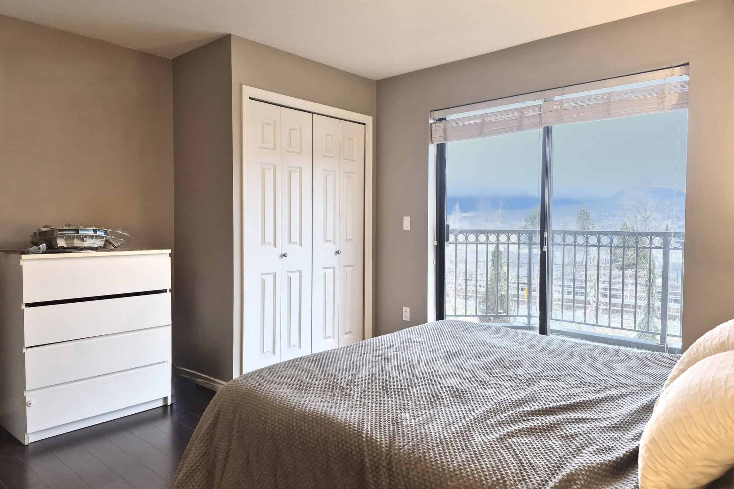 Modern bedroom with large balcony overlooking city and mountains, featuring neutral decor, closet, and natural light, ideal for Vancouver real estate and property investment.