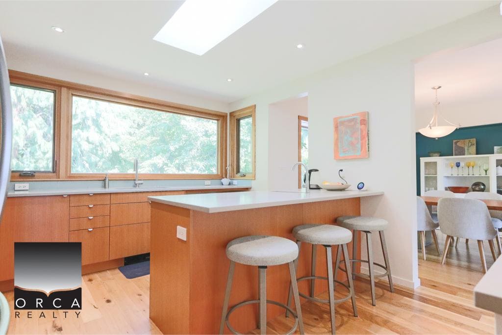 Vibrant kitchen with large windows showcasing lush greenery, wooden cabinetry, and modern bar stools, perfect for Vancouver Island homebuyers seeking stylish and functional spaces.