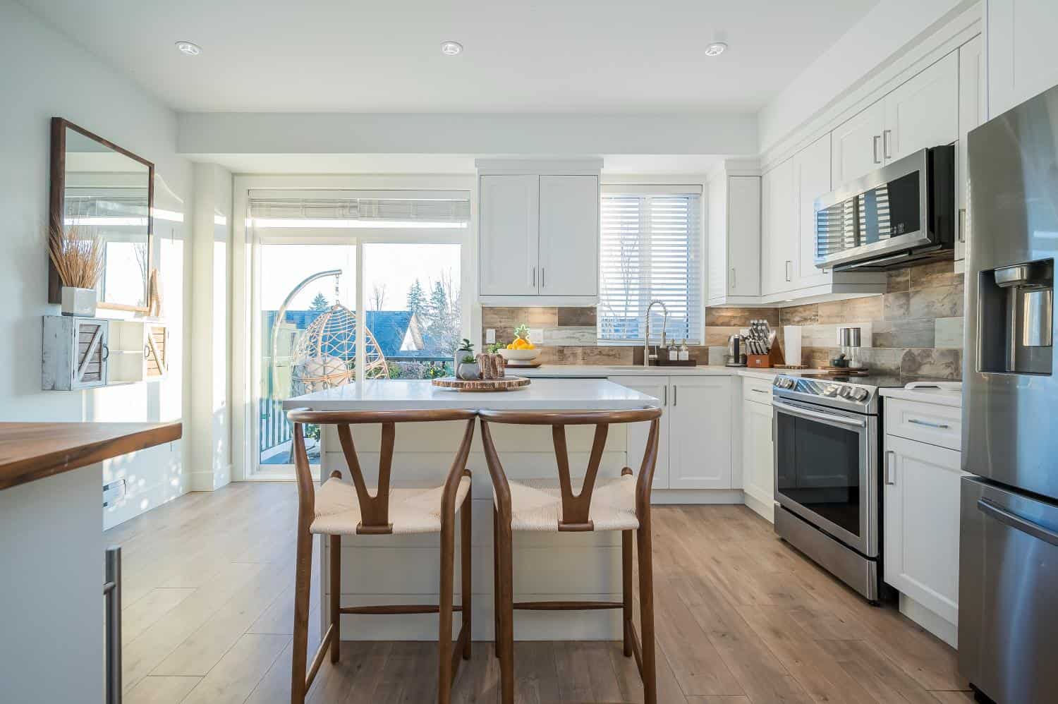 Bright modern kitchen with white cabinetry, stainless steel appliances, and a spacious island, featuring natural light from large windows and sliding door leading to outdoor balcony.