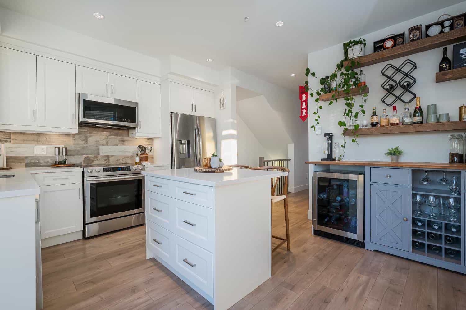 Bright modern kitchen with white cabinetry, stainless steel appliances, and a bar area featuring wooden shelves, wine storage, and decorative accents, showcasing Vancouver Island real estate.