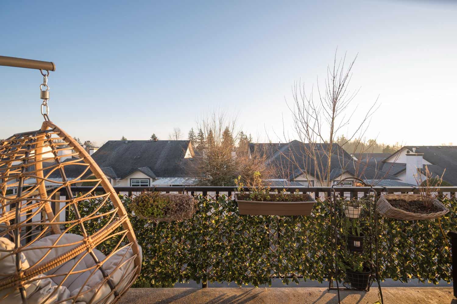 Cozy outdoor balcony with potted plants and woven hanging chair, overlooking a suburban neighborhood with rooftops and trees, perfect for relaxed living and outdoor entertaining.