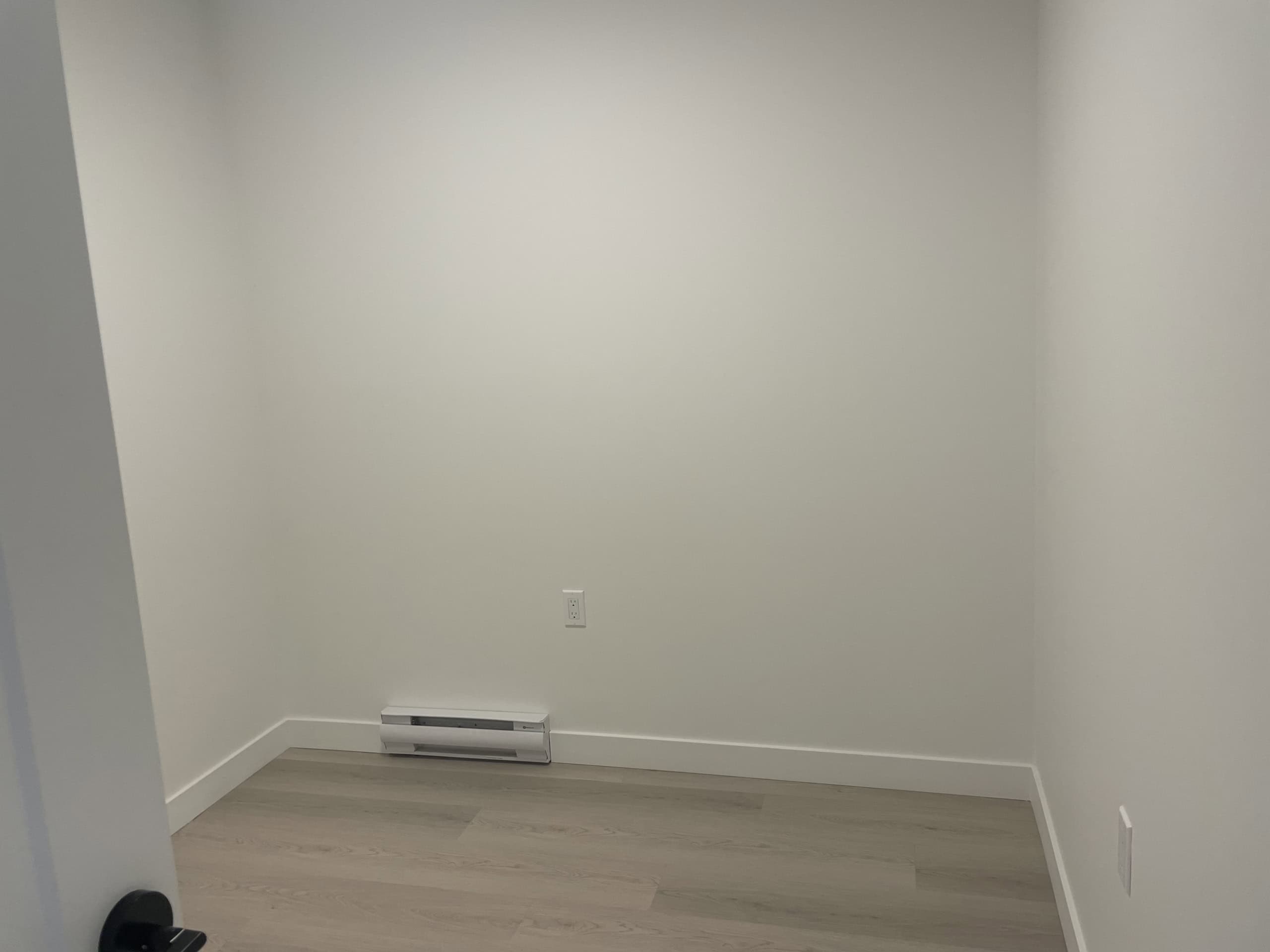 Bright, empty room with white walls, light wood flooring, and baseboard heating, showcasing modern interior design for potential real estate buyers or renters seeking Vancouver area homes.