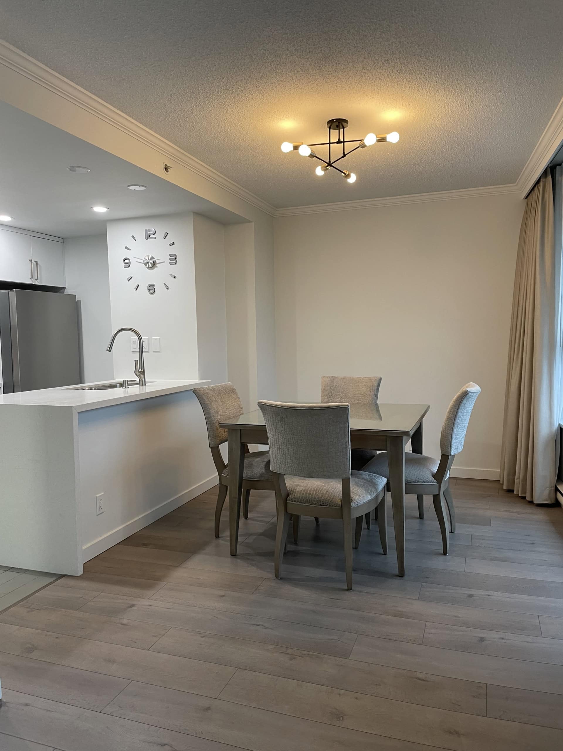 Bright modern dining area with a stylish chandelier, adjacent to a sleek kitchen with clock and stainless steel appliances, ideal for Vancouver real estate listings.