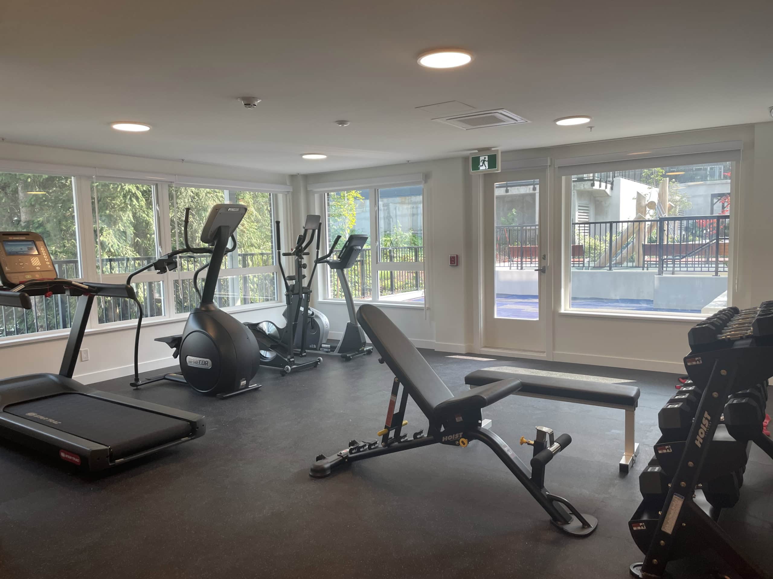 Modern fitness room with cardio equipment, free weights, and weight benches, bright with natural light and large windows overlooking outdoor amenities, perfect for residents' active lifestyles.