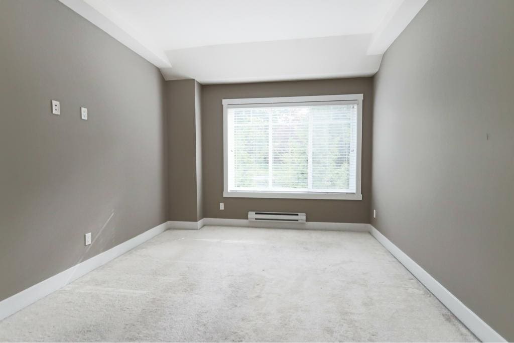 Bright empty bedroom with large window, grey walls, white trim, and carpet flooring, showcasing a modern living space ideal for Vancouver Island real estate.